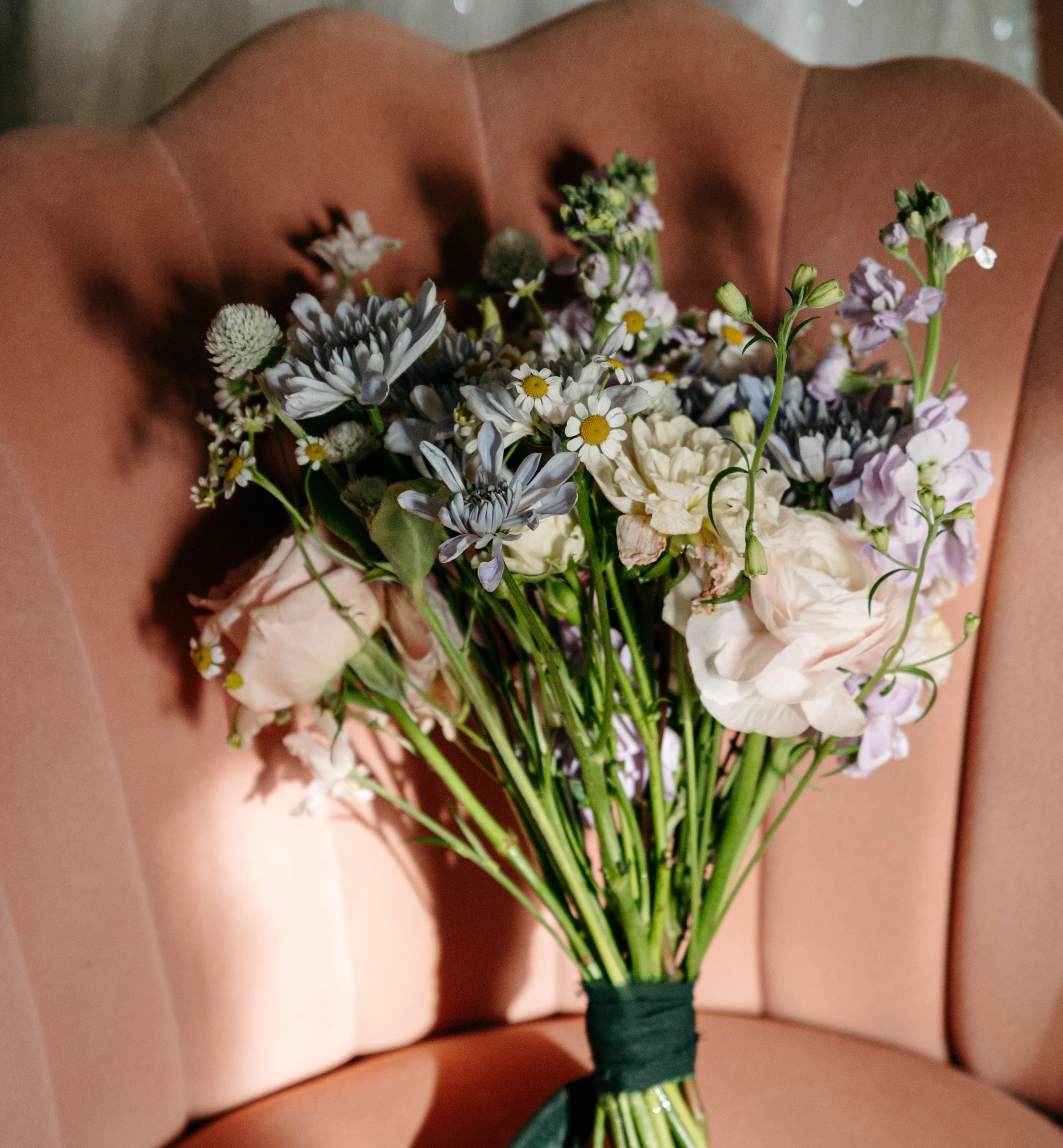 Hand-tied bouquet of soft white, blush, lavender, and pale blue flowers resting on a peach velvet chair in warm sunlight