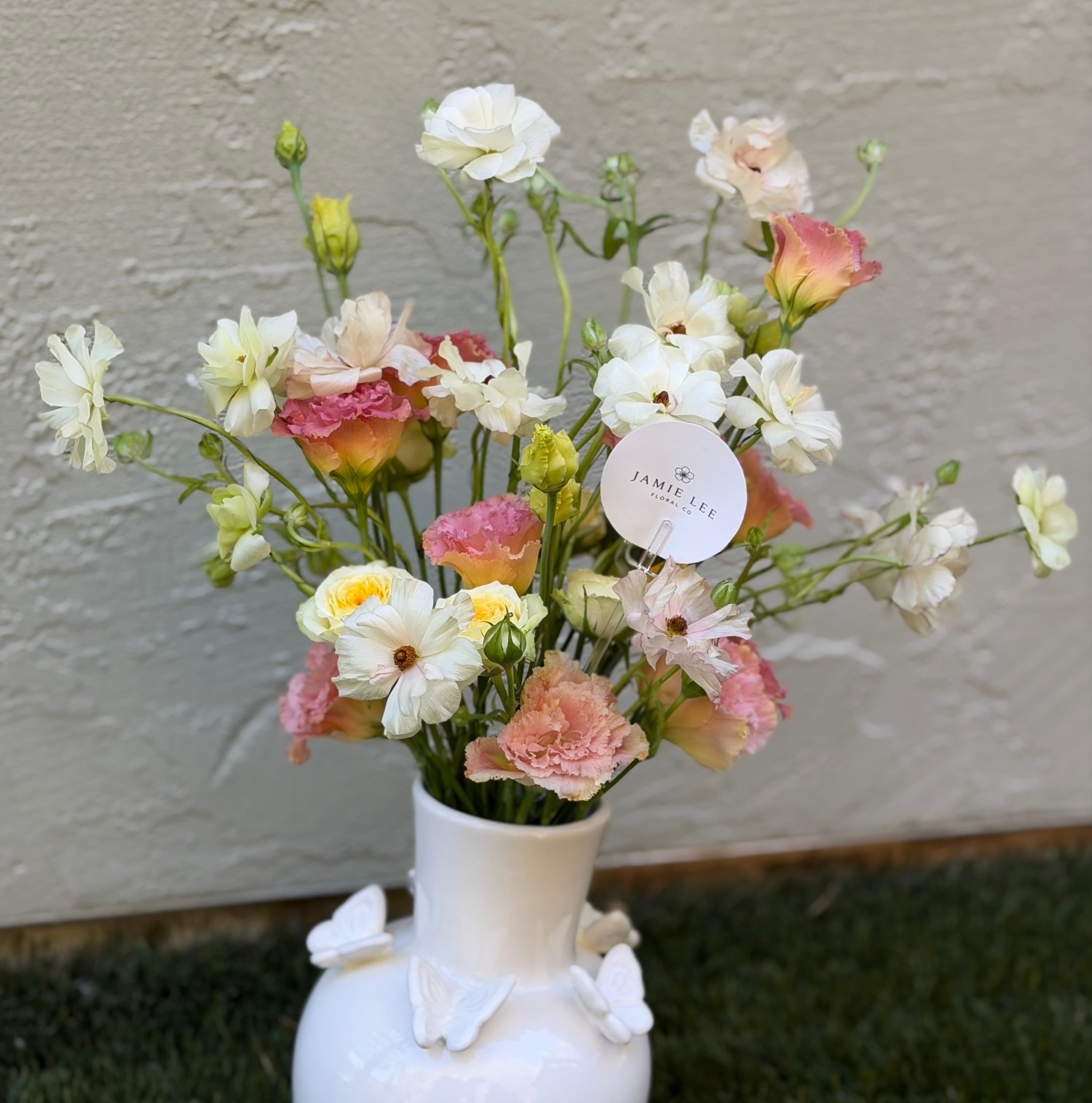 Pastel floral arrangement with white ranunculus, pink lisianthus, and roses in a white butterfly vase by Jamie Lee Floral Co.